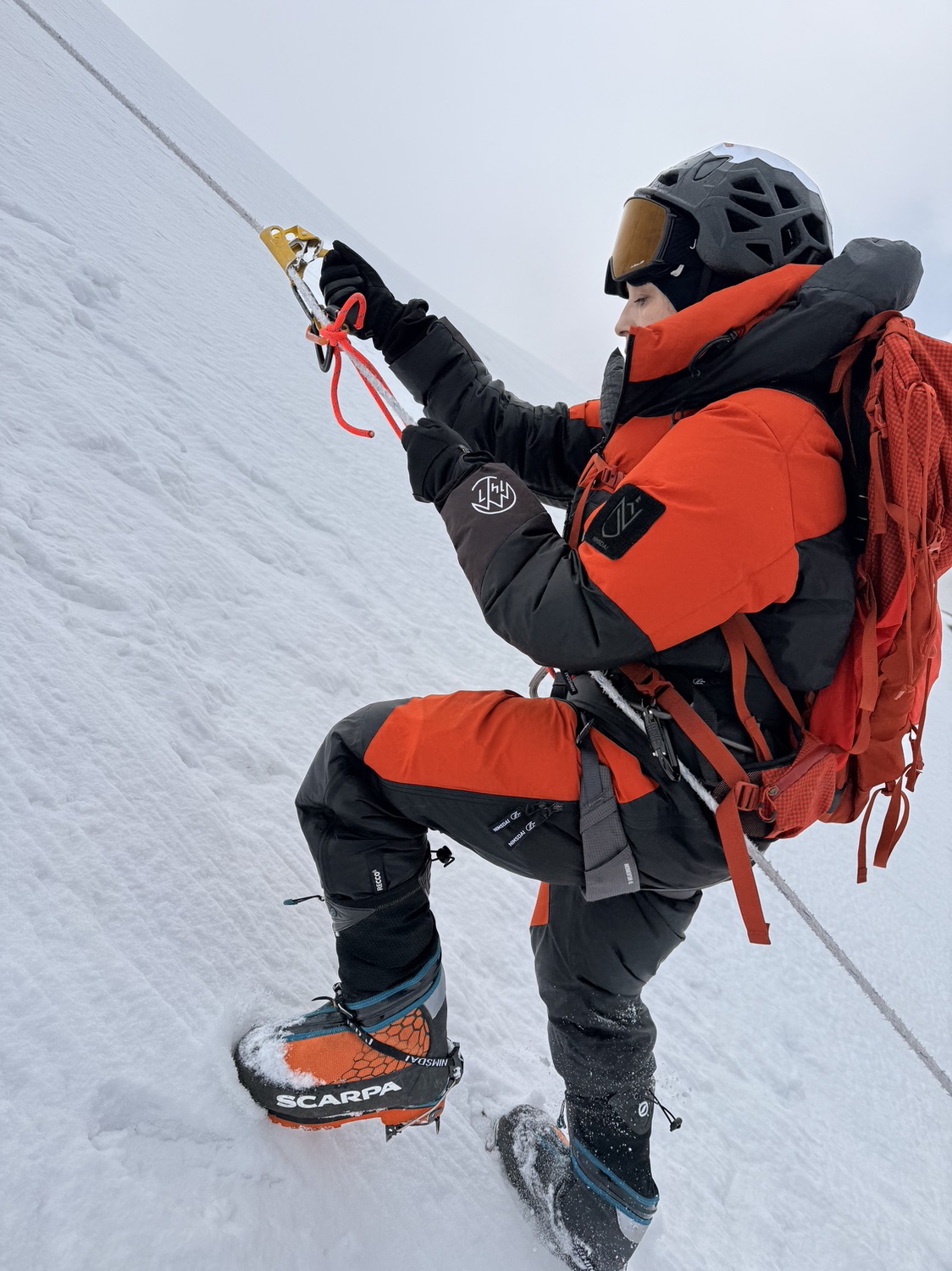 Adriana Aristizábal climbing a fixed rope on the snow slopes of Lobuche Peak, in helmet, goggles and orange expedition suit