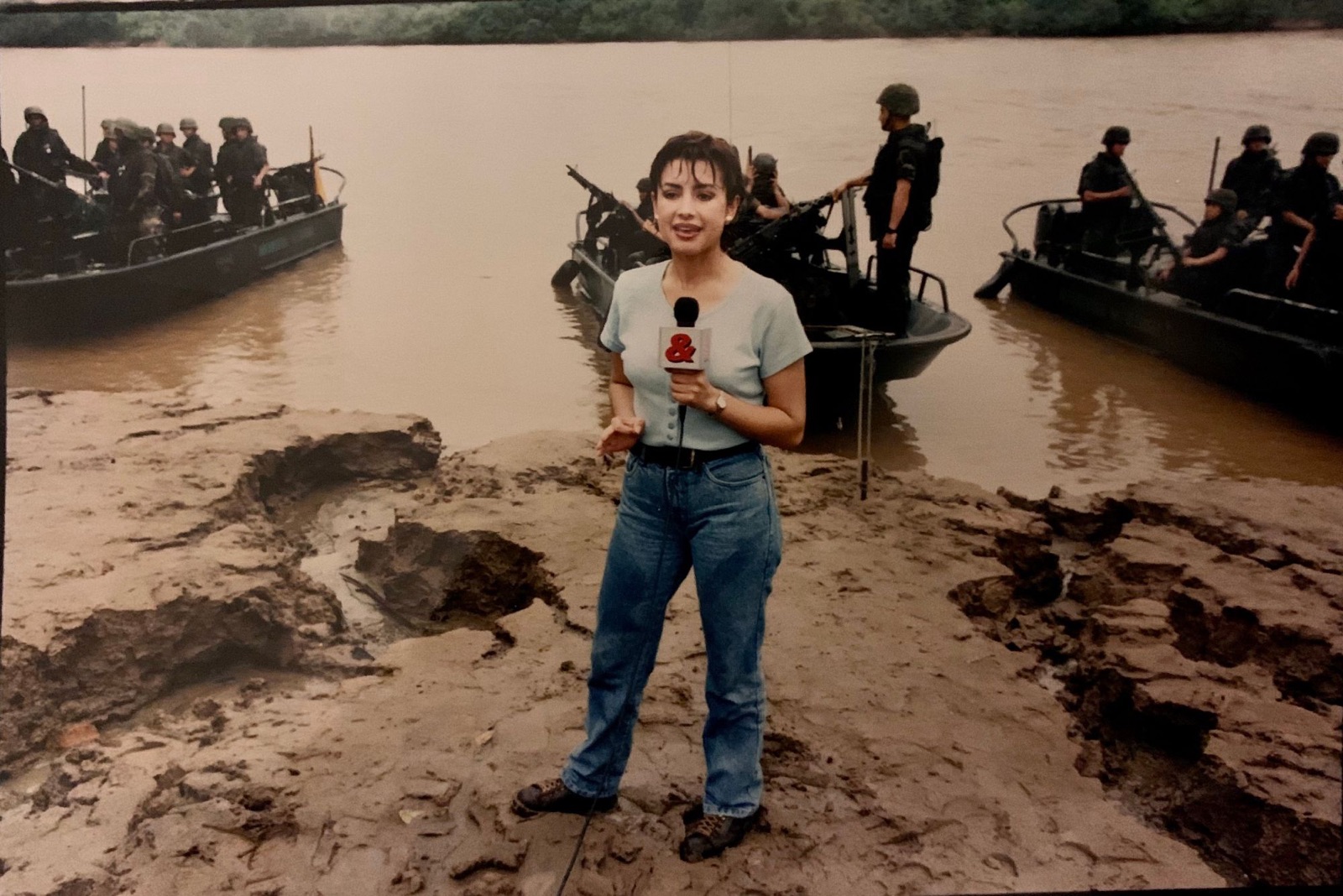Adriana Aristizábal reporting for RCN Televisión on a riverbank in Colombia, with armed military patrol boats behind her
